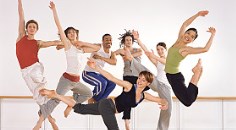 six ballet dancers jumping in midair, one female ballerina standing on one leg in front of them