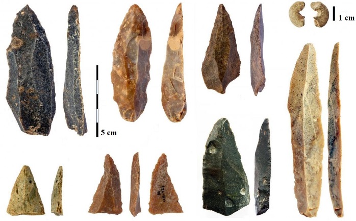 Eight stone blades and fragments, gray and tan in color, ranging from 1 cm to approximately 10 cm long, shown in face and profile views; most are shaped with a sharp point at one end; one is a stone bead split in half; scale bars of 5 cm and 1 cm are shown.