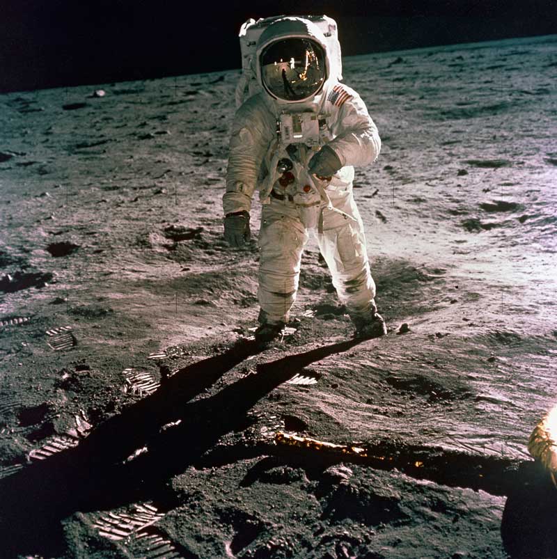 Buzz Aldrin in a spacesuit on the Moon, with the black of space in the sky and the rugged, gray, lunar landscape in the background and foreground