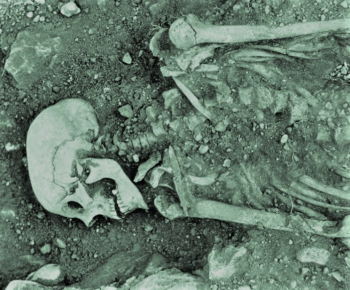 Partially excavated human skeleton; skull, upper torso, and arm bones can be seen in the dirt and rocks
