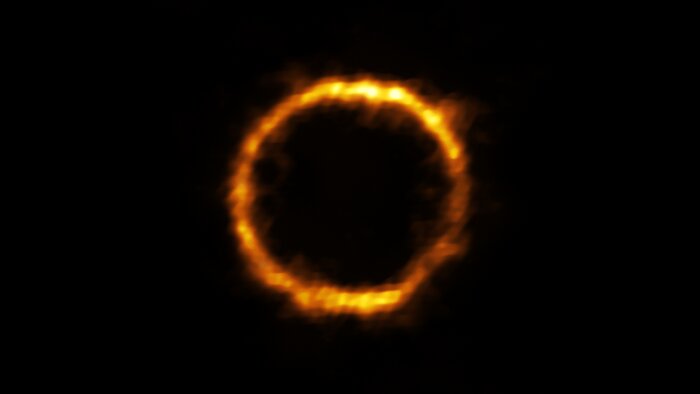 An orange ring of varying brightness against a black background. The ring is actually a gravitationally lensed, very distant galaxy