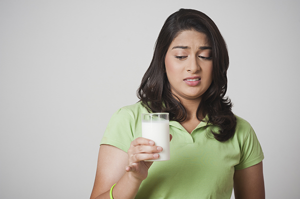 a woman wearing a light green polo shirt and standing against a white background, seen from the torso up, and holding a clear glass of milk. The woman has an expression of disgust on their face, indicated by narrowed eyes, elevated eyebrows, and curled upper lip.