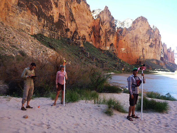 three researchers standing on a Grand Canyon sandbar holding surveying instruments and recording data