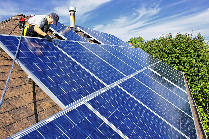 man at top on roof installing blue solar panels with trees visible in the background