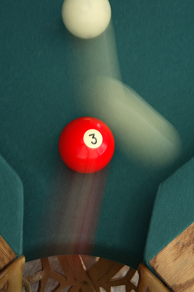 motion capture image of white cue ball, blurred, striking a red numbered solid billiard ball against the blue-green felt backdrop of a billiards table 